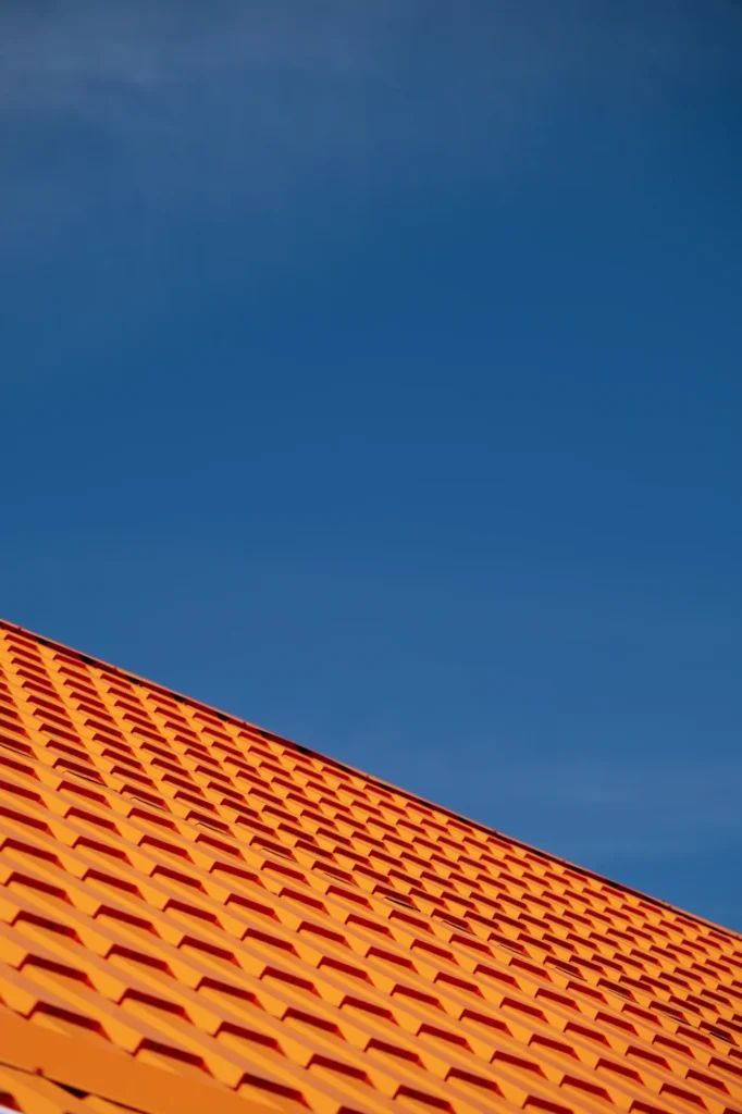 Toit orange vif et ciel bleu profond : contraste architectural. Toiture métallique inclinée orange vif au motif géométrique répétitif. L'architecture moderne contraste avec le ciel bleu profond.